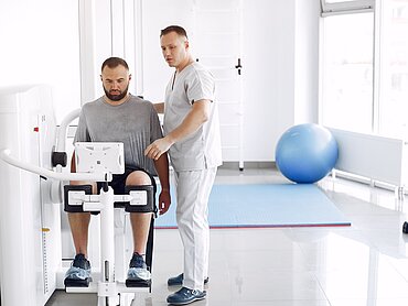 Reha Ein Mann sitzt auf einem physiotherapeutischen Gerät. Daneben steht ein Mann, der ihm etwas erklärt.