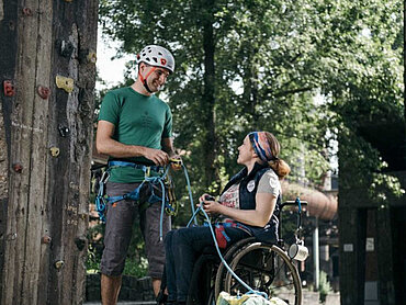Ein Mann mit Helm und eine Frau im Rollstuhl vor einer Kletterwand.