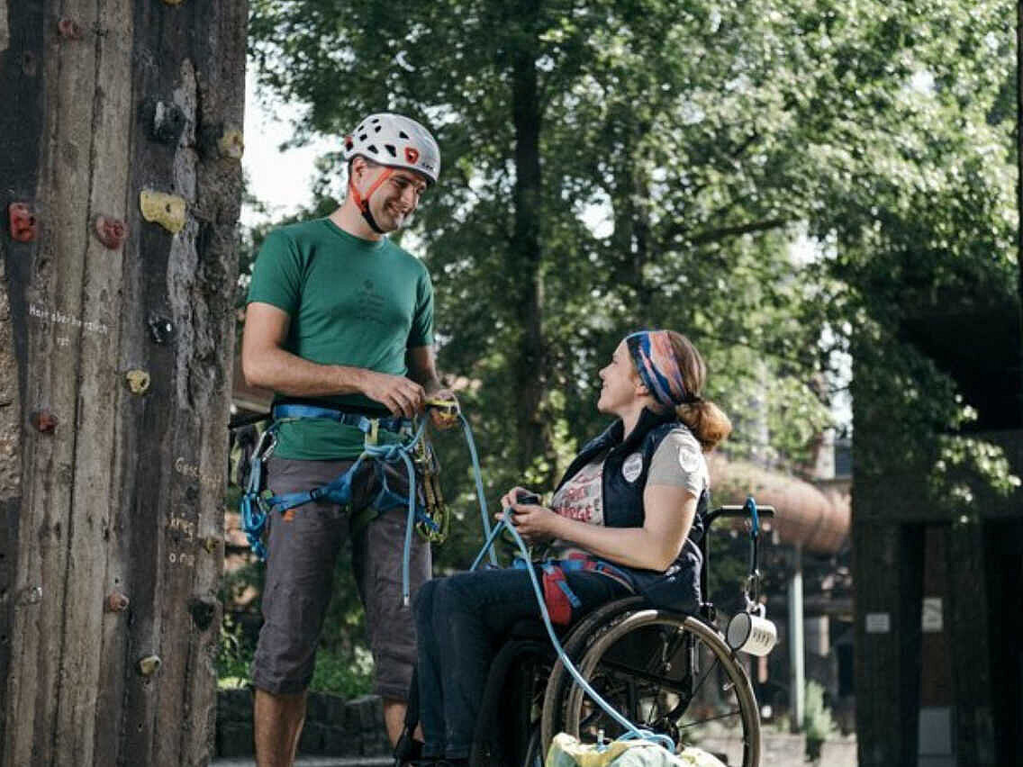 Ein Mann mit Helm und eine Frau im Rollstuhl vor einer Kletterwand.