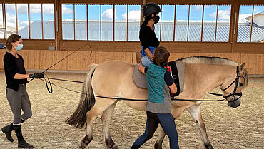Eine Frau auf einem Pferd, das im Schritttempo läuft. Sie wird von einer anderen Frau gestützt, die neben dem Pferd läuft. Eine weitere Frau führt das Pferd.tzt  