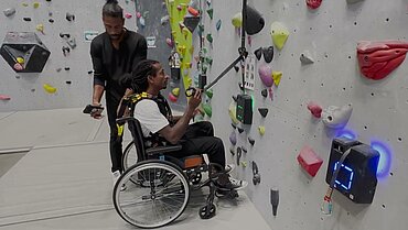 Zwei Menschen, davon einer im Rollstuhl, vor einer Kletterwand.