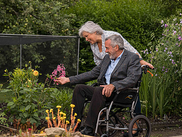 Eine Frau schiebt einen Mann im Rollstuhl durch einen Garten.