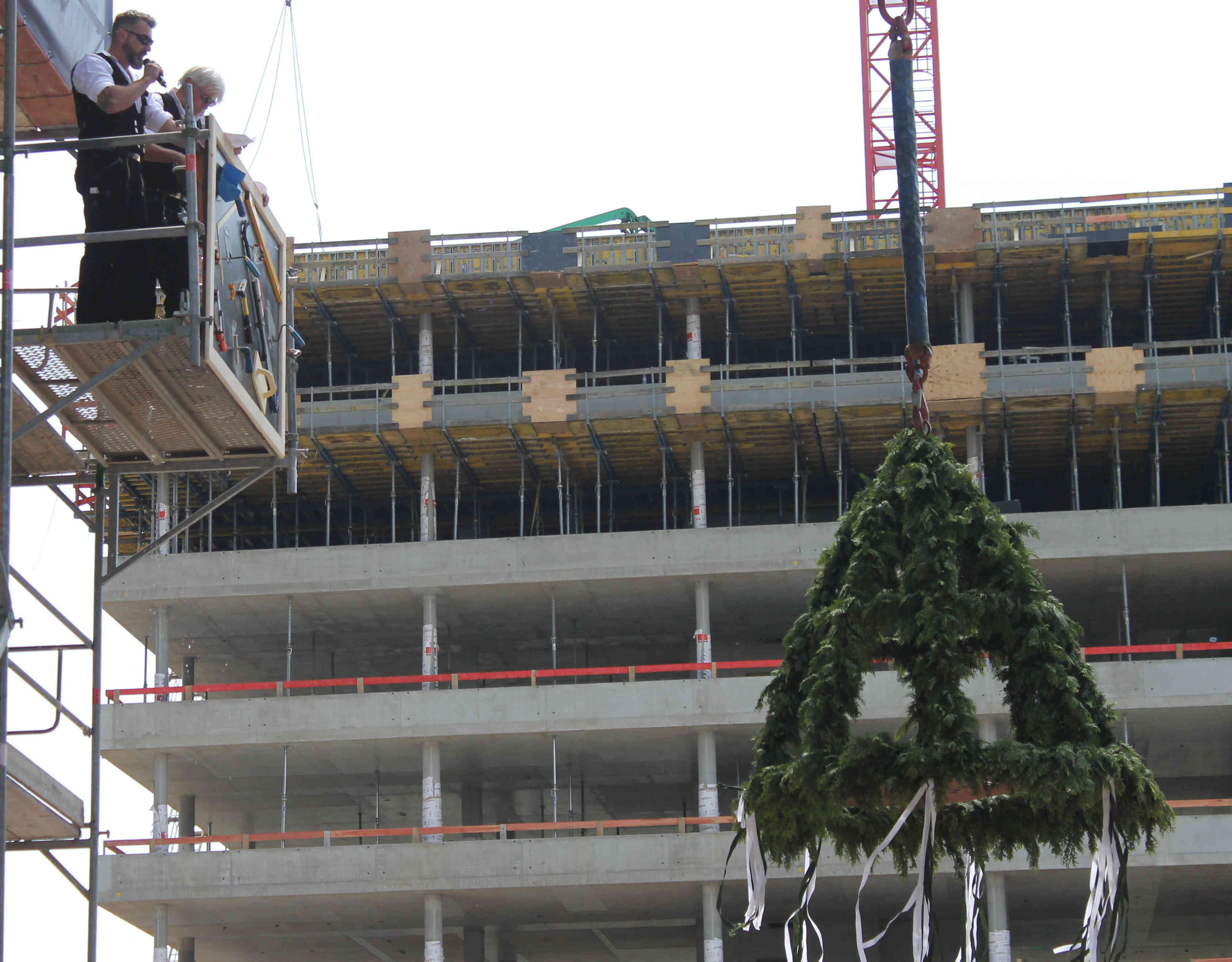 Zwei Zimmermänner auf einem Baugerüst. Vor ihnen ein Richtbaum an einem Kran.